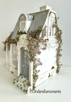 a white doll house with pink flowers growing on the roof and windows, in front of a white background