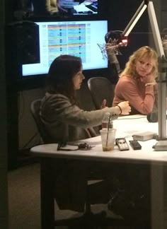 two women sitting at a table with microphones in front of them, talking to each other