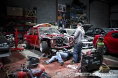 two men working on a car in a garage with other cars and tools around them