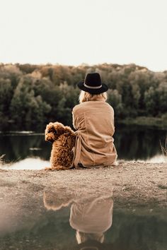 a woman sitting on the ground next to a body of water with a dog in her lap