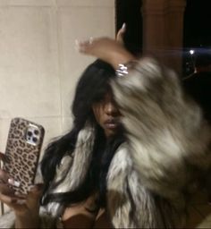 a woman with long hair holding a cell phone in her hand and wearing a fur coat