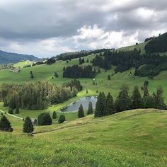 a lush green hillside covered in trees next to a lake surrounded by mountains and clouds