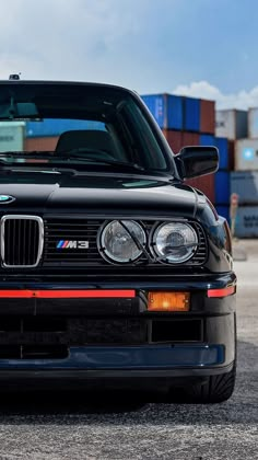 the front end of a black bmw car parked in a lot with shipping containers behind it