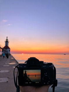 a person holding up a camera to take a photo on the water at sunset with a lighthouse in the background