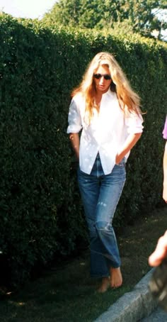 two women are walking down the sidewalk in front of some hedges and bushes, one is wearing sunglasses