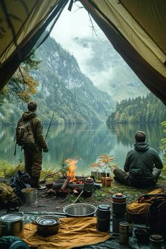 two men sitting at a campfire in front of a lake with mountains and trees