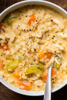 a white bowl filled with chicken and vegetable soup on top of a wooden table next to a spoon