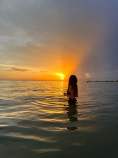 a woman is sitting in the water watching the sun go down over the ocean with her back turned to the camera