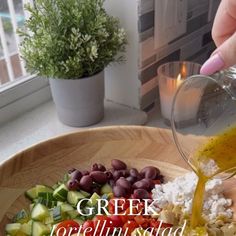 a person pouring olives, cucumbers, and other ingredients into a bowl