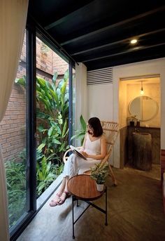 a woman sitting on a chair reading a book in front of a large window with plants outside