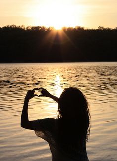 a woman is standing in the water making a heart shape with her hands at sunset