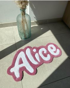 a pink and white door mat with the word alice on it next to a vase