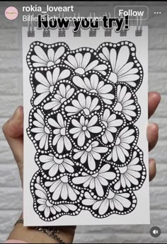 a hand holding up a drawing with black and white flowers on the paper, which reads'now you try '