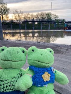 two green stuffed animals sitting on top of a wooden bench next to a lake and bridge