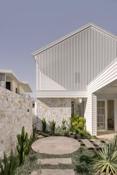 a house with white walls and stone walkway leading up to the front door is surrounded by greenery