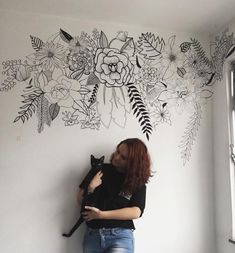 a woman holding a cat in front of a wall with flowers and leaves on it