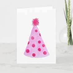 a card with a pink and red party hat on the front, sitting next to a green plant