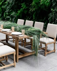 an outdoor dining table with white chairs and green plants on the wall in the background