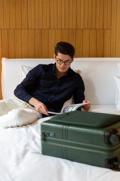 a man sitting on a bed reading a book next to a green suitcase and white sheets