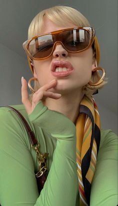 a woman wearing sunglasses and a scarf with her hand on her chin posing for the camera