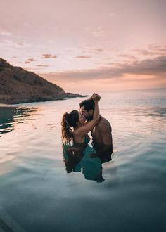 a man and woman are kissing in the water at sunset with mountains in the background