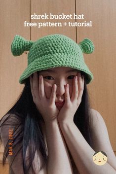 a girl wearing a green crocheted hat covering her face with both hands while looking at the camera