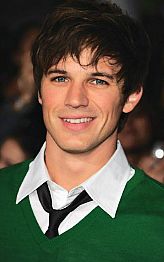 a young man wearing a green sweater and black tie with people in the background taking pictures