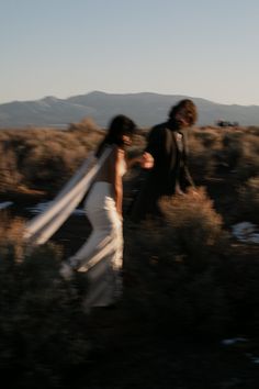 a bride and groom are walking through the brush in their wedding attire, with mountains in the background