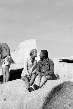 three men sitting on top of large rocks in the desert, one is talking to another man