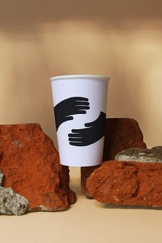 a coffee cup with two hands on it sitting next to some rocks and red clay