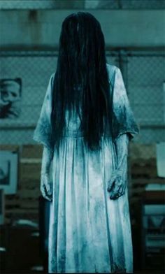 a woman with long hair standing in a dark room, wearing a white dress and holding her hands behind her back