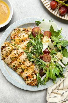 a white plate topped with chicken and salad next to a bowl of dressing on top of a table