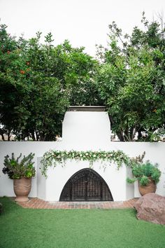 an outdoor fireplace surrounded by potted plants and trees in front of a white wall