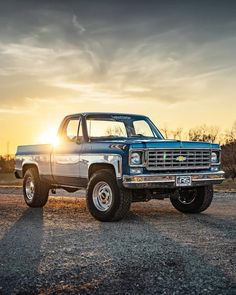 a blue pickup truck parked on the side of a road with the sun setting in the background