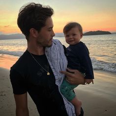 a man holding a baby on the beach at sunset