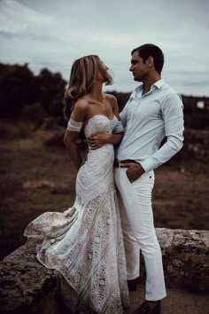 a man and woman standing next to each other on top of a rocky hill with the ocean in the background