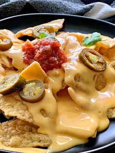 a black plate topped with nachos covered in cheese sauce and pickle slices