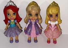 three little princess dolls standing next to each other on a white surface with one wearing a tiara