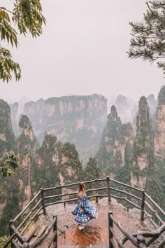 a woman sitting on top of a wooden bench next to trees and mountains in the background