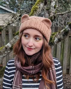 a woman wearing a knitted hat and scarf in front of a fence with trees