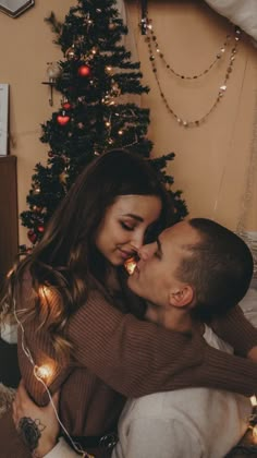 a man and woman are kissing in front of a christmas tree with lights on it