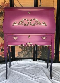 a pink painted dresser with gold trim and ornate designs on the top, in front of a floral wallpapered background