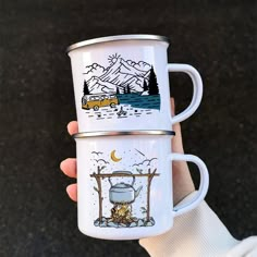 two white coffee mugs with drawings on them are held in front of the camera