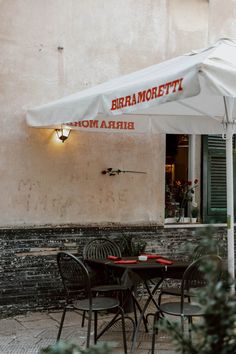 tables and chairs under an umbrella on the side of a building with flowers growing outside