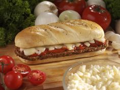 a sandwich with meat and cheese on a cutting board next to tomatoes, onions, garlic and parsley