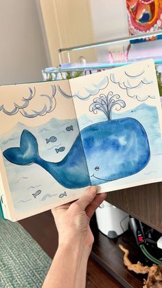 a person holding up an open book with watercolors on it and a drawing of a whale