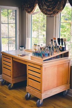 a wooden desk topped with lots of paint and brushes on top of it next to a window