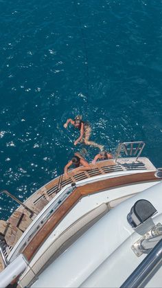 two people hanging out on the deck of a sailboat in the blue water while another person is fishing