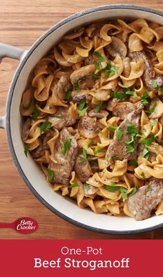 a pot filled with pasta and meat on top of a wooden table next to silverware