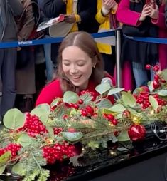 a woman smiles as she sits in front of a table with holly garlands and red berries